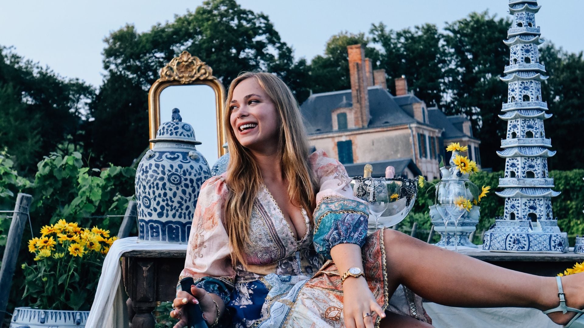 Woman sitting at a table in a chateau vineyard near Reims surrounded by Chinese blue and white porcelain towers and jars, enjoying Taittinger champagne with sunflowers and mirror decor